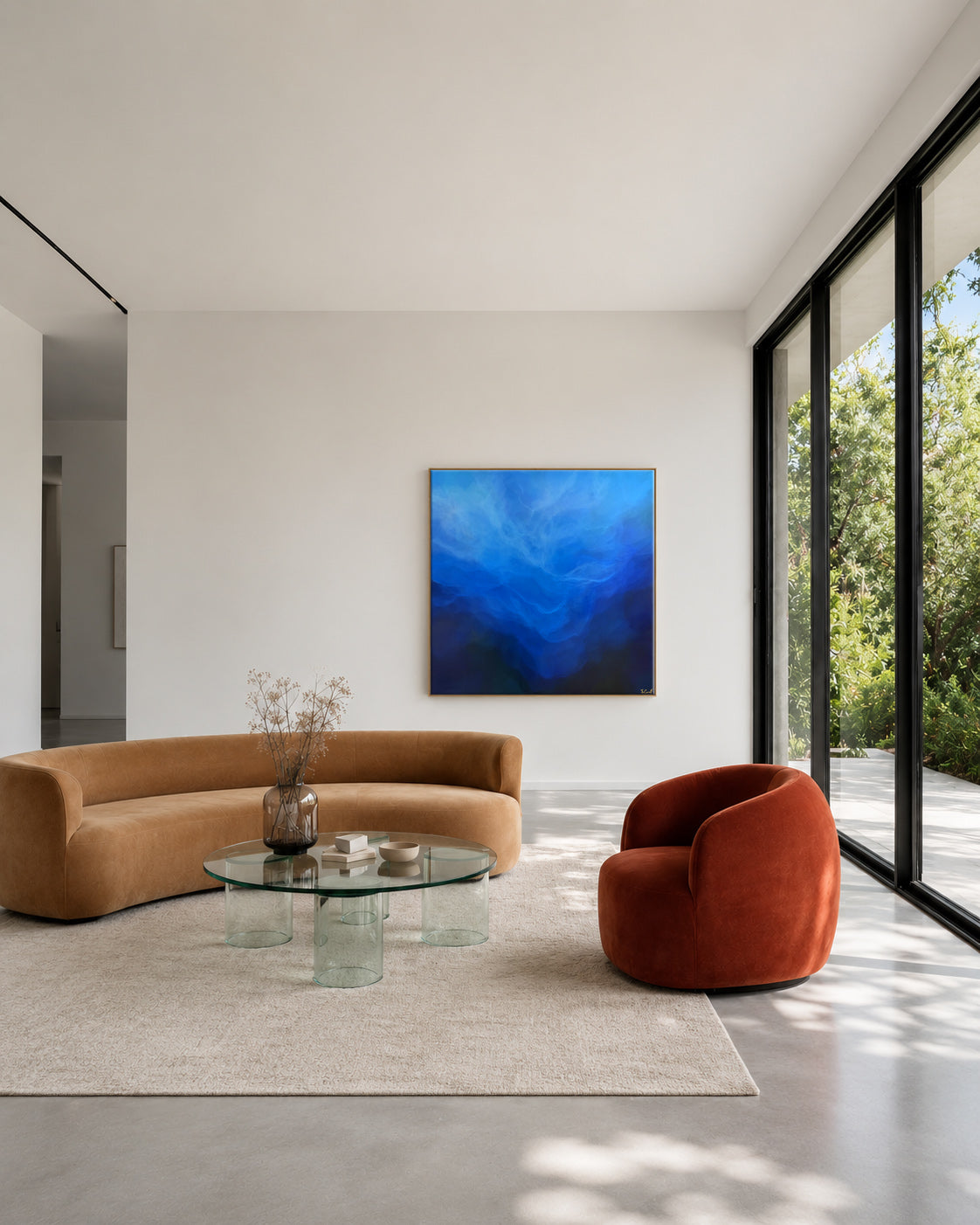 A modern living room interior with a curved tan sofa, a terracotta armchair, a glass coffee table, and a large abstract blue painting centered on a white wall.
